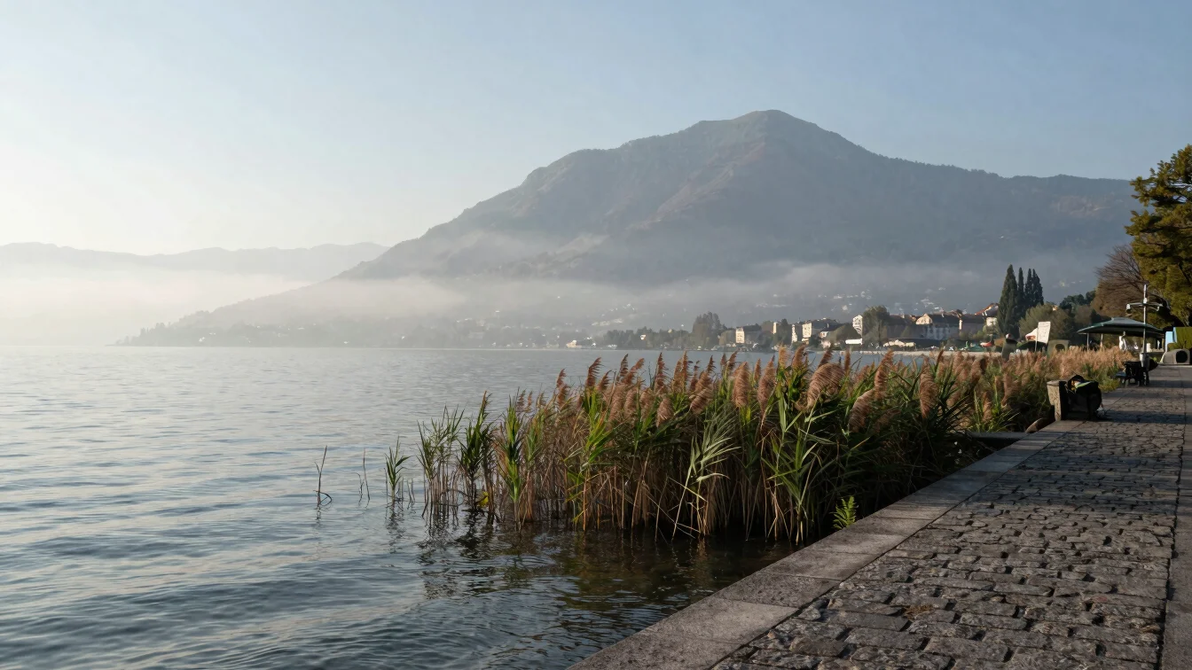 Calm early-morning view of Lake Lugano shoreline with soft mist, a stone promenade and reeds, muted blue-green palette,